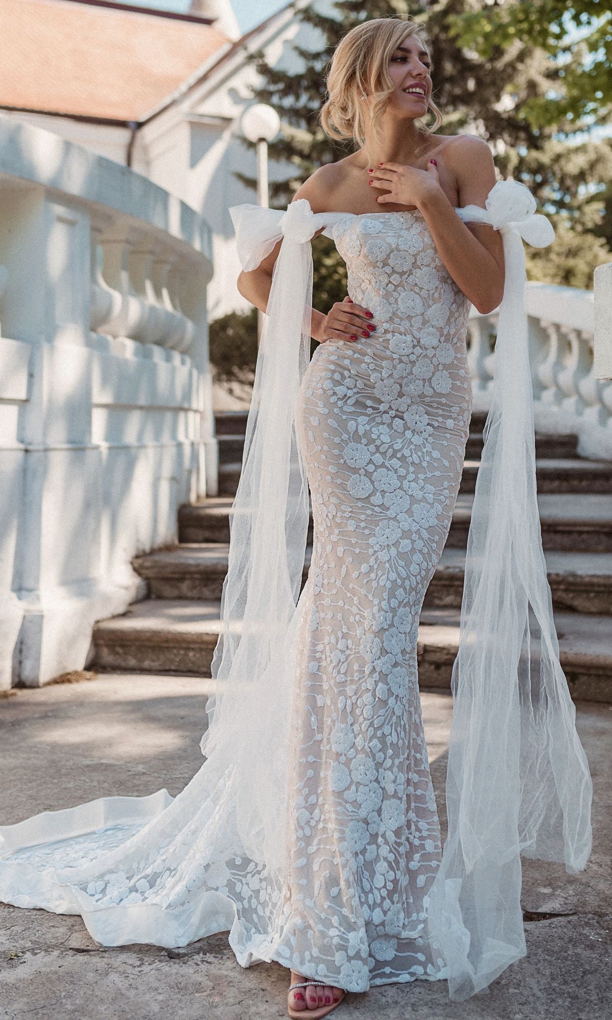 A woman wearing the Strapless White Wedding Gown: Tina Holly BB010, featuring a fitted mermaid silhouette and sheer lace accents, stands smiling on outdoor stone steps with one hand to her chest.